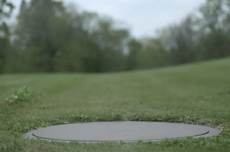 A plain round septic tank access lid set flush in a mown green lawn in rural Iowa