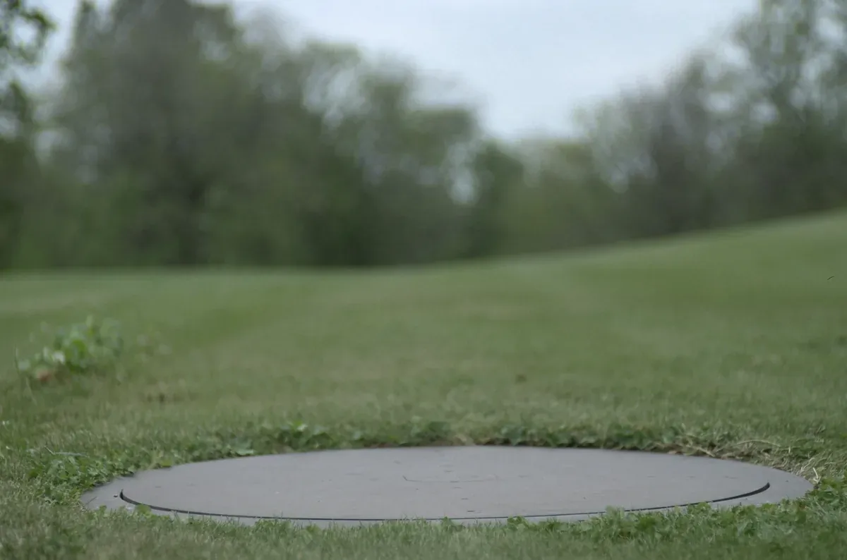 A plain round septic tank access lid set flush in a mown green lawn in rural Iowa
